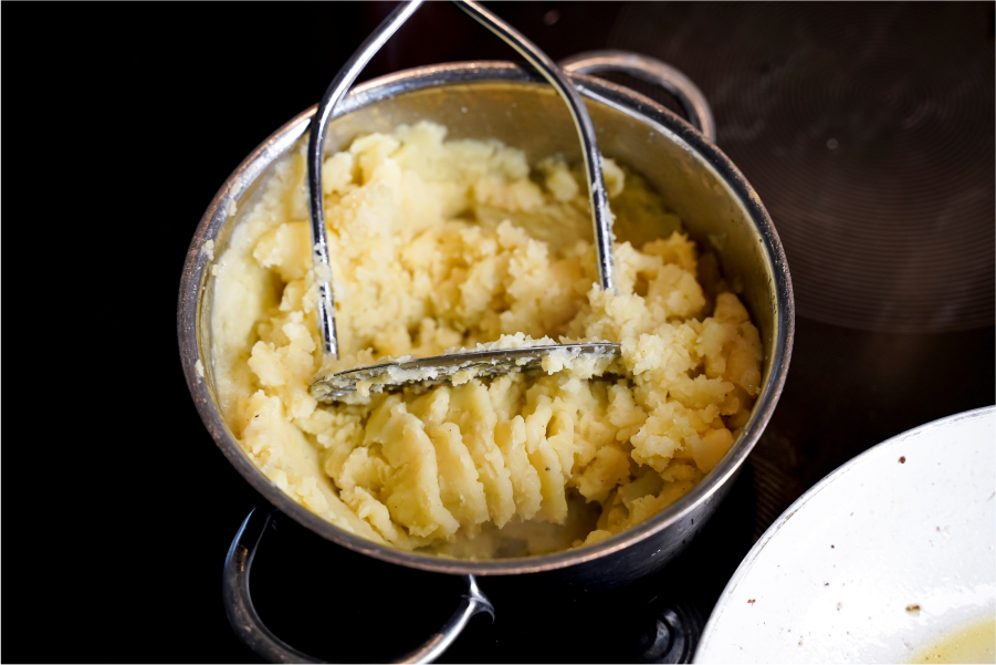 A stainless steel saucepan containing mashed potatoes and a potato masher sitting upon a glass cooktop