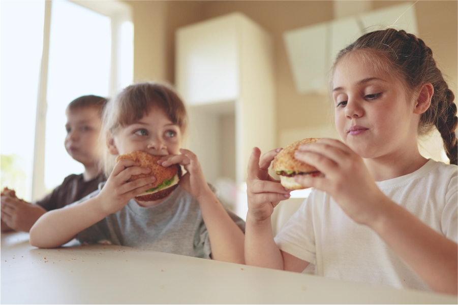 Three children seated at a table eating filled rolls