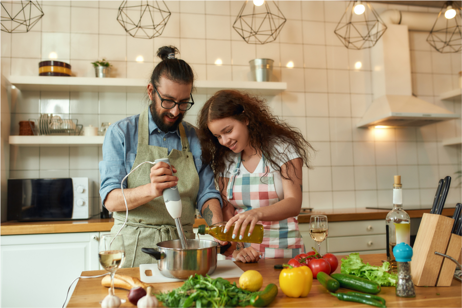 A young couple preparing a meal in a well lit kitchen, the man is using a stick blender in a stainless steel pot as the woman pours olive oil into it. On the bench are an assortment of fresh vegetables.