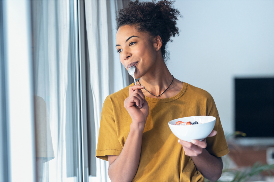 A woman eating a bowl of yoghurt with fruit as she looks wistfully out of a window