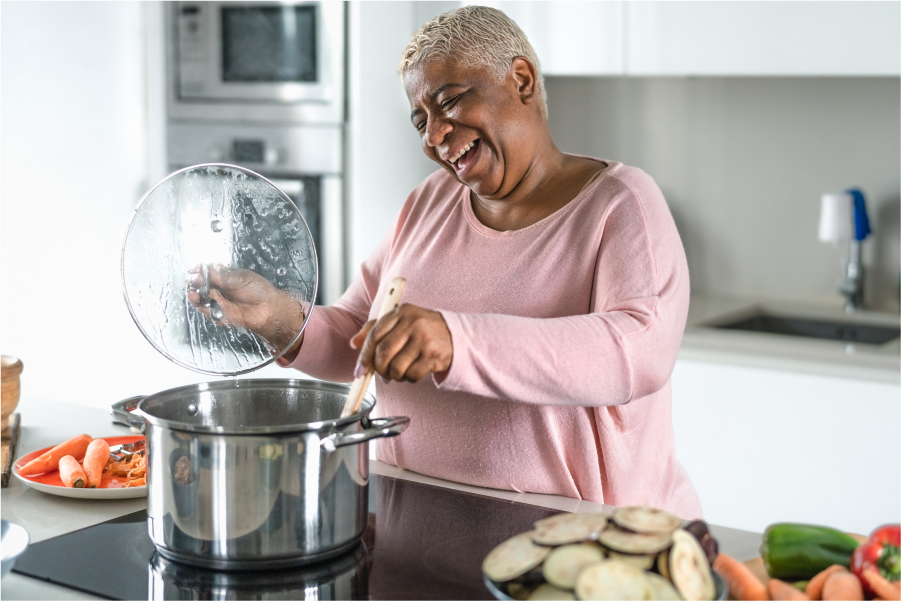 A woman in a modern kitchen stirring a stainless steel stock put with an assortment of sliced vegetables on the bench next to her