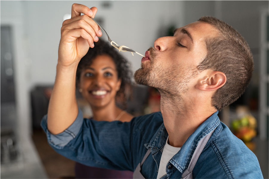 A man eating a noodle from a fork as a woman in the background looks on in amusement