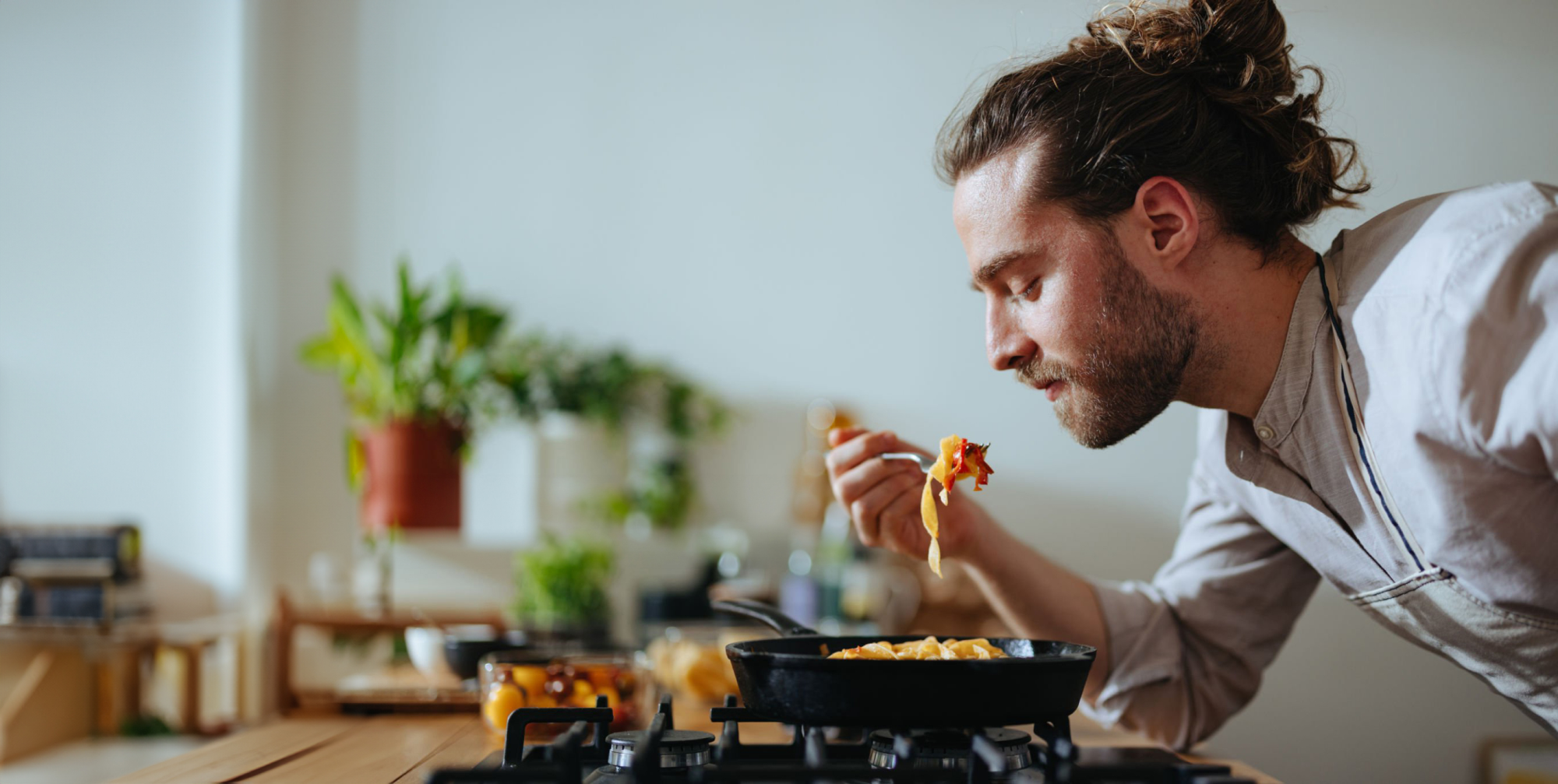 A long-haired man eating pasta from a cast iron skillet on a gas cooktop