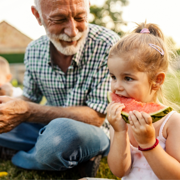 A young girl eating a slide of watermelon seated outside as an elderly gentleman looks on in amusement